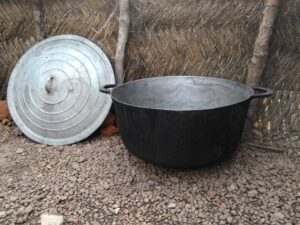Grande marmite en métal avec son couvercle posé à côté, utilisée pour la cuisson de repas en grande quantité dans un village africain.