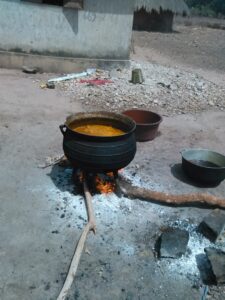 Un cuisinier prépare de l'huile rouge africaine dans une marmite sur un feu ouvert, entouré d'ingrédients traditionnels.