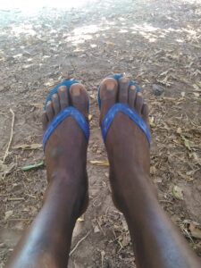 Un homme assis sous un arbre imposant, les pieds étendus, dans un paysage africain vaste et tranquille. L'ombre de l'arbre lui offre un refuge contre la chaleur du soleil.
