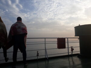 Plusieurs hommes debout sur un bateau à l'aube, leurs silhouettes se détachant contre un ciel lumineux. L'eau calme est enveloppée de brume, créant une atmosphère paisible et mystérieuse.