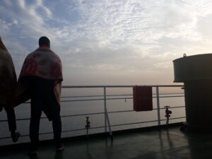 Plusieurs hommes debout sur un bateau, leurs silhouettes se détachant contre le ciel lumineux de l'aube, avec des couleurs chaudes et une brume légère sur l'eau.