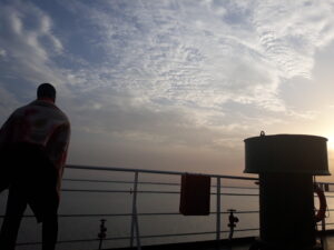 Silhouettes de plusieurs hommes debout sur un bateau, regardant l'horizon à l'aube en Afrique. La lumière douce du matin éclaire l'eau calme, avec un voile léger de brume.