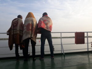 Silhouettes de plusieurs hommes debout sur un bateau, face au lever du soleil en Afrique. L'atmosphère est calme, baignée de lumière douce et de couleurs chaudes, avec un léger voile de brume sur l'eau.
