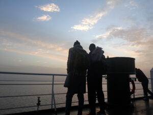 Silhouettes de plusieurs hommes debout sur un bateau au lever du soleil en Afrique, avec des couleurs chaudes et une lumière douce du matin. L'horizon est légèrement brumeux, créant une atmosphère paisible et intime.