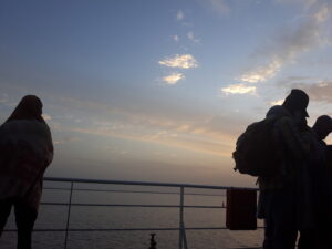 Silhouette d'un homme debout sur un bateau au lever du soleil en Afrique, avec des couleurs chaudes et une lumière douce du matin. L'horizon est légèrement brumeux, et l'atmosphère est calme et sereine.