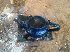 Image d'une petite cafetière posée sur un petit fourneau, chauffé par un feu de charbon, dans un contexte africain